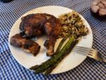 blue and white checkered tablecloth with a white plate, on the plate is a grilled BBQ chicken leg quarter, grilled asparagus and an orzo pasta salad